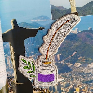 Puede incluir: Parche bordado con una pluma, un tintero con tinta morada y una ramita de hojas. El parche tiene un borde plateado y está colocado sobre un fondo de paisaje urbano y estatua.