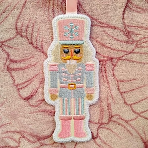 May include: A fabric Christmas ornament shaped like a nutcracker soldier. The ornament features a pink hat with a snowflake, a light blue uniform with pink accents, and a tan face with a yellow mustache. A pink ribbon is attached at the top.