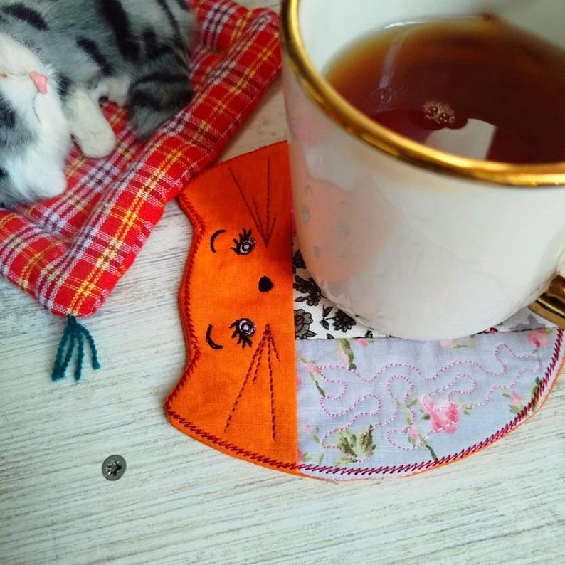 Pu&ograve; includere: Sottobicchiere in tessuto a forma di gatto arancione e blu con occhi e baffi ricamati. Il sottobicchiere &egrave; su una superficie di legno bianco con una tazza di t&egrave; sopra.