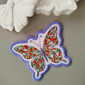 May include: Embroidered butterfly patch with a colourful striped wing design in red, yellow, green, and blue. The patch has a light purple border and is on a grey surface. A white butterfly-shaped mould is in the background.