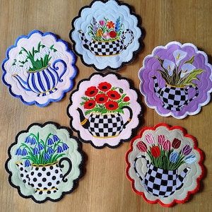 May include: A collection of seven decorative, embroidered coasters. Each coaster features a teapot design filled with various flowers. The teapots have different patterns, including checkered and polka dots, and are set against scalloped backgrounds in various colors.