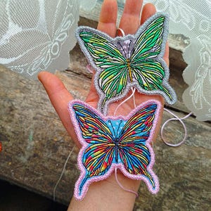 Könnte beinhalten: Zwei schmetterlingsförmige, gestickte Aufnäher. Einer hat grüne und gelbe Flügel mit einem grauen Rand. Der andere hat regenbogenfarbene Flügel mit einem rosa Rand. Beide haben filigrane Fadendetails und befinden sich auf hellem Hintergrund.