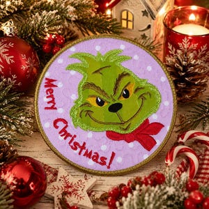 May include: A circular embroidered patch featuring a green character with yellow eyes and a red scarf on a light purple background with white polka dots. The text Merry Christmas! is stitched in red thread along the left side of the patch.