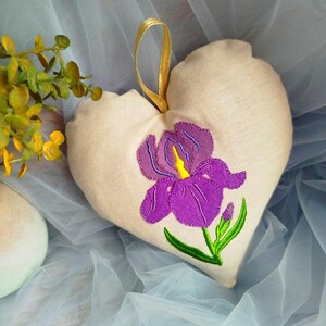 May include: A heart-shaped beige fabric pillow with a purple embroidered iris flower. The pillow has a gold ribbon loop for hanging.