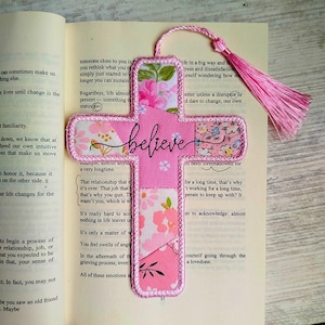 May include: A pink cross-shaped bookmark with the word "believe" in cursive. The bookmark is made of fabric with floral patterns and a pink tassel. It is placed on an open book.