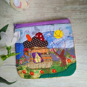 May include: A colourful fabric zipper pouch featuring a mushroom house design. The house has a brown roof with white polka dots, a purple door, and a yellow window. The background is a patchwork of blue, green, and brown fabrics with embroidered flowers and trees.