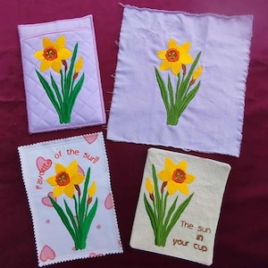 May include: Four embroidered items featuring a yellow daffodil with green leaves and stems. One item is a lavender quilted pouch. Another is a square of lavender fabric. The third has the text "Favorite of the sun" and hearts. The fourth has the text "The sun in your cup".
