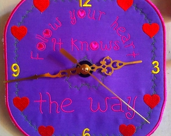 May include: Purple fabric clock with a gold clock face and hands. The clock face has the words "Follow your heart it knows the way" embroidered in pink. The numbers 12, 9, 6, and 3 are embroidered in yellow. The clock is surrounded by a pink border with red hearts.
