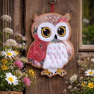 May include: A decorative owl wall hanging with large, expressive eyes. The owl features a quilted design with floral patterns in red and brown, a white fluffy chest with pink accents, and yellow feet. It hangs from a red string.