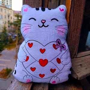 May include: A light purple cat-shaped plush toy with a heart-shaped design on its chest. The heart is decorated with red hearts and a purple bow. The cat has black whiskers, a pink nose, and pink cheeks.