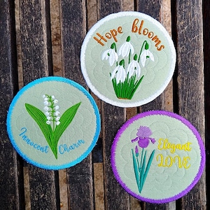 May include: Three embroidered circular patches with floral designs and inspirational phrases. One features snowdrop flowers and the words "Hope Blooms." Another shows lily of the valley with "Innocent Charm." The third has an iris and "Elegant Love."