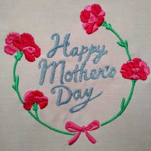 Peut inclure: Broderie représentant une couronne de oeillets roses avec des tiges et des feuilles vertes. La couronne entoure le texte "Happy Mother's Day" en lettres cursives bleues.