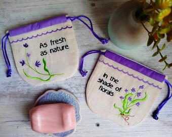 May include: Two small, beige fabric drawstring bags with purple trim. The bags are embroidered with floral designs and text. The bag on the left says "As fresh as nature" and the bag on the right says "In the shade of florals".