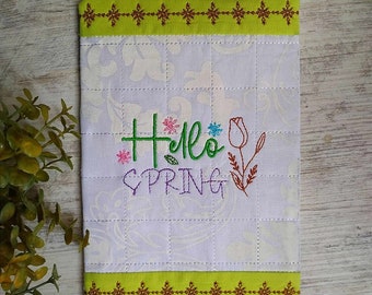 May include: A white fabric pouch with a green and brown border. The pouch has a quilted design and features embroidered text that says "Hello Spring" with a flower design.