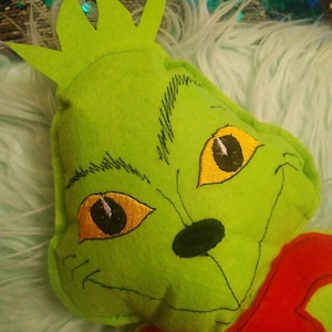Könnte beinhalten: Grünes Plüschtier aus Filz mit einem gestickten Gesicht, gelben Augen und einer schwarzen Nase. Das Spielzeug hat eine rote Schleife aus Filz um den Hals.