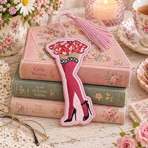 May include: A fabric bookmark shaped like a pair of legs wearing a red floral skirt, pink thigh-high stockings with black and white checkered trim, and black high-heeled shoes. The bookmark features a light pink tassel attached to the top.