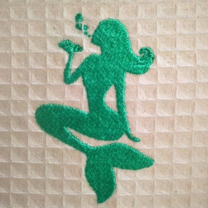May include: Embroidered green mermaid design on a white towel. The mermaid is sitting with her tail curled behind her and blowing bubbles.