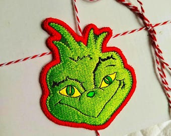 May include: Embroidered green Grinch face patch with a red border, featuring yellow eyes and a smiling expression. The patch is attached to a white package with red and white twine, ideal for Christmas gifts.