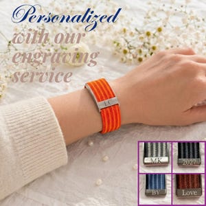 May include: A vibrant orange and red striped fabric bracelet featuring a silver-toned metal clasp engraved with the letters LC. Additional examples show variations with grey, black, blue, and red straps engraved with MK, 2027, BY, and Love.