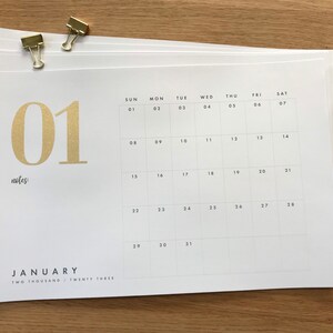 May include: A stack of white paper calendars for January 2023, held together with gold binder clips. The calendar features a minimalist design with gold lettering and numbers. The month and year are printed at the bottom.