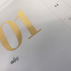 May include: Close-up of a white calendar page with gold foil numbers and the word "notes:" in black script. The number "01" is prominently displayed, with the numbers "08" and "15" visible in the background.