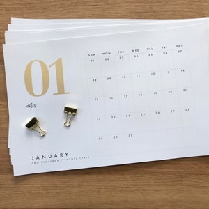 May include: A white paper calendar for January 2023 with gold-colored text and accents. The calendar is open to the first month, with the number "01" prominently displayed. Two gold binder clips and a black and white notebook are also visible.