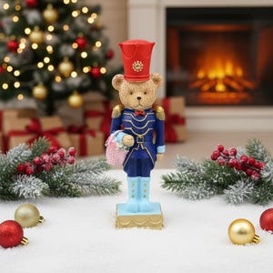 Può includere: Una statuetta di orso in piedi vestito da soldato schiaccianoci, con un cappello rosso, un'uniforme blu scuro con finiture dorate e stivali azzurri. L'orso tiene un piccolo cestino rosa pieno di fiori e poggia su una base rettangolare dorata.