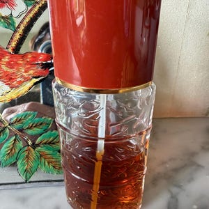 Può includere: Un decanter cilindrico in vetro con un motivo a foglie in rilievo sulla superficie esterna, riempito con un liquido color ambra. Il decanter presenta un alto coperchio rosso tinta unita con bordo dorato e una cannuccia in plastica bianca.