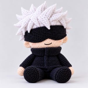May include: A handcrafted crochet doll featuring spiky white hair, a black blindfold covering the eyes, and a high-collared black outfit. The doll has a light peach-colored complexion, a simple stitched mouth, and a seated posture.