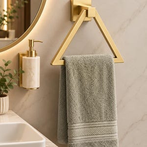May include: A triangular gold metal towel ring mounted on a wall, holding a folded sage green textured hand towel with a decorative hem. Beside it, a wall-mounted soap dispenser features a cylindrical marble body with gold metal hardware.