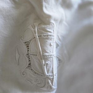 Peut inclure: Un textile en lin blanc orné d'un monogramme brodé ton sur ton. Le motif présente des volutes élaborées et des détails ajourés entourant les lettres centrales, mettant en valeur un travail d'aiguille raffiné sur un tissu tissé lisse.