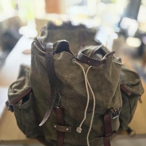 Könnte beinhalten: Ein olivgrüner Rucksack aus Segeltuch mit braunen Lederriemen und Metallschnallen, einem Kordelzugverschluss mit weißer Schnur sowie zwei Seitentaschen. Die Tasche hat eine strukturierte Stoffoberfläche mit sichtbaren Nähten und Metallbeschlägen.