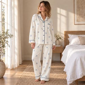 May include: A two-piece pajama set in off-white fabric featuring a repeating pattern of small blue bows. The long-sleeved button-down top has a notched collar and dark blue piping, while the matching long pants feature a straight-leg design.