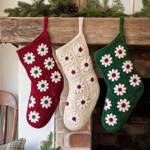 May include: Three crochet Christmas stockings hanging in a row. The left stocking is deep red, the center is cream, and the right is dark green. Each stocking features a repeating pattern of white daisy motifs with small colored centers.