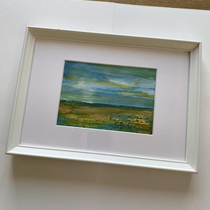 Könnte beinhalten: Ein gerahmtes Landschaftsgemälde mit einer texturierten Horizontlinie in Blau-, Grün- und Goldtönen. Das Kunstwerk befindet sich in einem schlichten, rechteckigen weißen Rahmen mit einem breiten weißen Passepartout, das einen klaren Kontrast bildet.