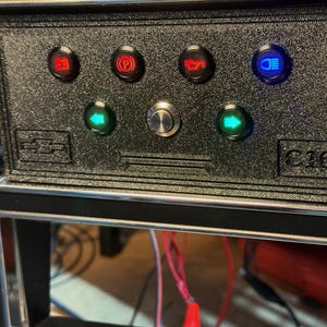 Può includere: Un pannello indicatore per cruscotto nero testurizzato per un veicolo Chevrolet C10. L'unità rettangolare presenta quattro spie LED nella fila superiore in rosso e blu, due frecce verdi per gli indicatori di direzione e un pulsante centrale.