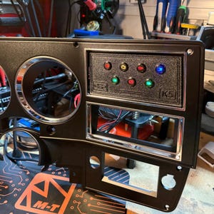 Puede incluir: Un panel de salpicadero de automóvil negro con dos grandes aberturas circulares para indicadores, una sección central rectangular con molduras cromadas y un panel de luces indicadoras con siete pequeñas luces de colores y el texto K5.