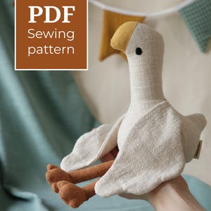 May include: A stuffed goose toy made of off-white textured fabric with a mustard yellow beak, brown legs, and a small black eye. A brown square graphic in the upper left corner contains the white text PDF Sewing pattern.