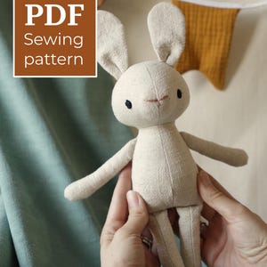 May include: A beige fabric rabbit doll with long ears, black embroidered eyes, and a pink stitched nose. A brown square graphic in the upper left corner contains the white text PDF Sewing pattern. The doll has a simple, stitched construction.