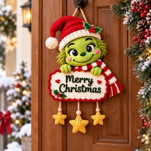 Könnte beinhalten: Ein gehäkelter Wandbehang mit einer grünen Figur, die eine rote Weihnachtsmannmütze und einen rot-weiß gestreiften Schal trägt. Die Figur hält ein weißes Schild mit dem grünen Text Merry Christmas und drei hängenden gelben Sternen.