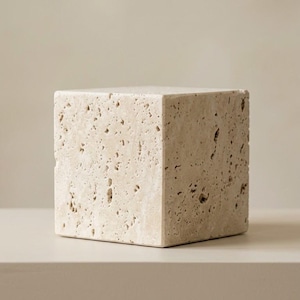 May include: A beige travertine stone cube with a porous, textured surface. The object is shaped as a perfect geometric cube, displaying natural variations in the stone's composition, including small pits and irregular indentations across its faces.