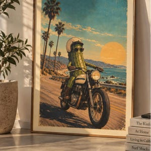 Puede incluir: Una lámina enmarcada que muestra un cactus verde con casco blanco y gafas de sol conduciendo una motocicleta por una carretera costera. La escena incluye palmeras, un gran sol amarillo y el océano bajo un cielo azul.