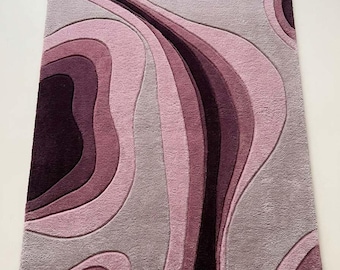 May include: A rectangular area rug featuring a layered, abstract topographic pattern in shades of deep plum, mauve, and light pink. The design consists of curved, flowing lines with varying pile heights, creating a textured surface. A small white tag with the text "SIR RUGS" is visible in the bottom right corner.