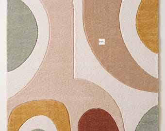 May include: A rectangular area rug featuring a tufted, abstract pattern of curved shapes in muted tones of sage green, terracotta orange, mustard yellow, beige, and cream. The design includes a small white tag with text in the center.