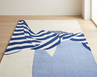 May include: A rectangular area rug featuring a cream-colored base with a large, solid light blue shape and a prominent graphic design of flowing, dark blue and white horizontal stripes that sweep across the surface in a wave-like pattern.