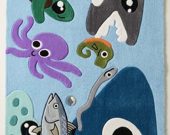 May include: A rectangular light blue rug featuring a tufted ocean scene with a grey shark, a purple octopus, a green sea turtle, a red crab, a yellow starfish, a small orange fish, a pink jellyfish, and a large fish. The text STE RUGS is visible.