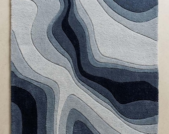 May include: A rectangular area rug featuring a layered, wavy pattern in shades of navy blue, slate gray, and light gray. The design mimics topographical lines with varying pile heights. A small white label in the corner reads STR RUGS.
