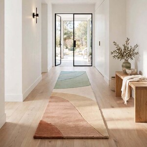 May include: A long rectangular runner rug featuring a series of overlapping curved shapes in muted tones of sage green, cream, beige, and terracotta, laid out on a light-colored wooden floor in a bright hallway with white walls.