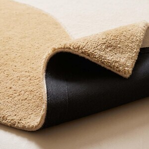 May include: A plush area rug featuring a curved color-blocked design in shades of beige and cream. The rug is folded over to reveal a textured, tufted top surface and a solid black, non-slip backing material on the underside of the fabric.