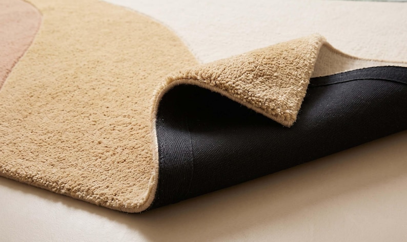 May include: A plush area rug featuring a curved color-blocked design in shades of beige and cream. The rug is folded over to reveal a textured, tufted top surface and a solid black, non-slip backing material on the underside of the fabric.